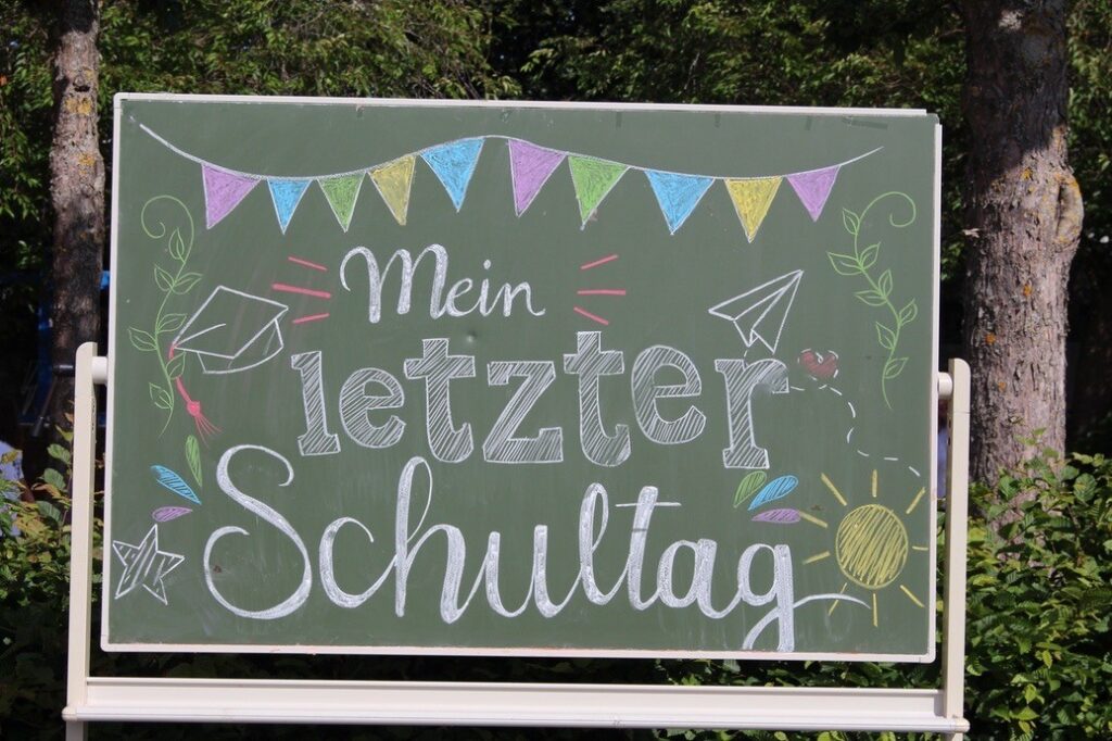 Eine Kreidetafel im Freien mit der bunten Aufschrift „Mein letzter Schultag“, verziert mit gemalten Wimpeln, einer Sonne und einem Doktorhut.
