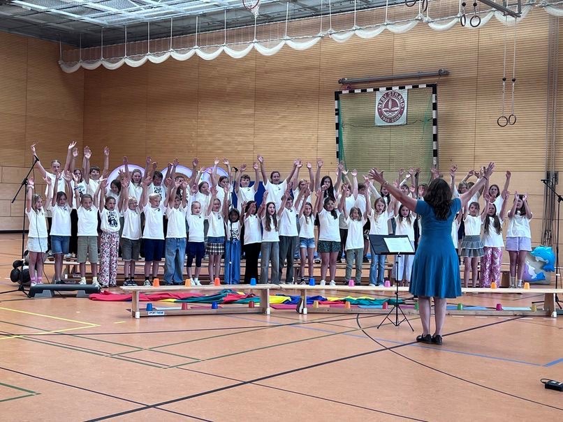 Ein großer Kinderchor steht auf Holzbänken in einer Turnhalle und singt mit erhobenen Armen, dirigiert von der Chorleiterin Uli von Welt