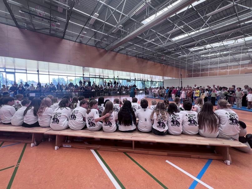 Kinder der 4. Klasse der GGS Timmendorfer Strand in einheitlichen T-Shirts sitzen auf einer Turnbank und schauen bei einer Schulveranstaltung in einer großen Sporthalle zu.